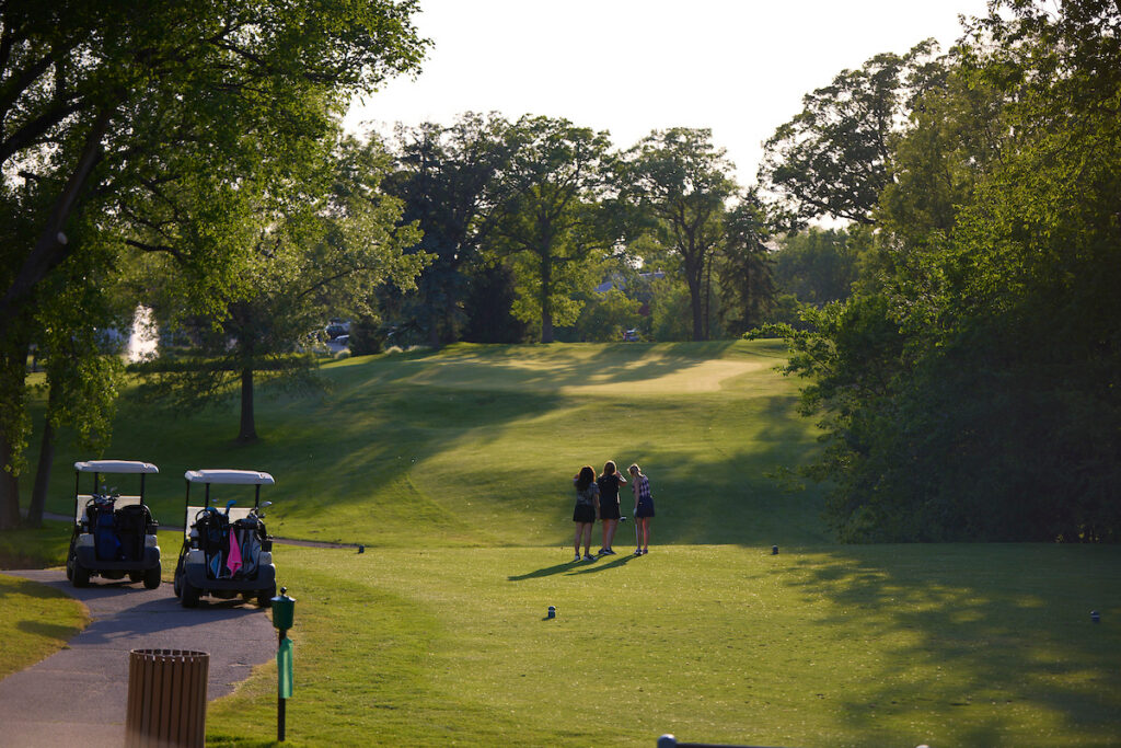 3 ladies examine the shot from the tee box looking toward the elevated green in the distance at dusk.
